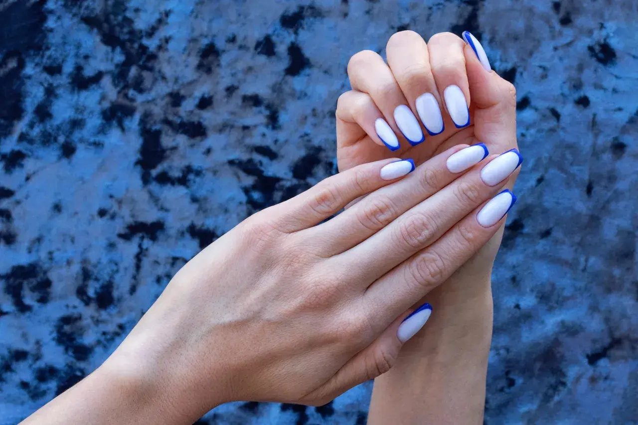 Eleganckie, białe paznokcie z granatowym french manicure, prezentowane na tle niebieskiego, aksamitnego materiału.